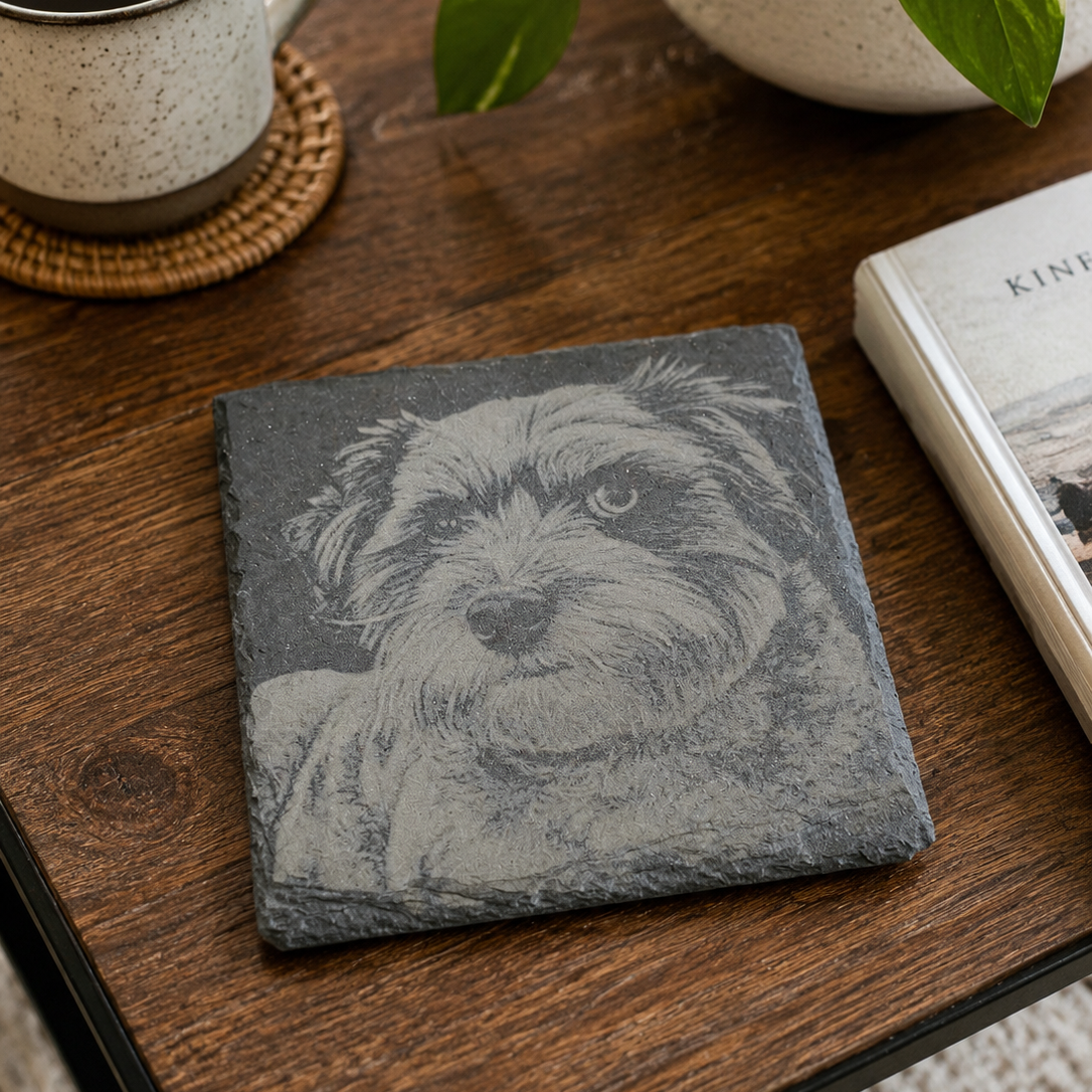 Slate coaster with dog illustration on a wooden table with a cup of coffee and plant.